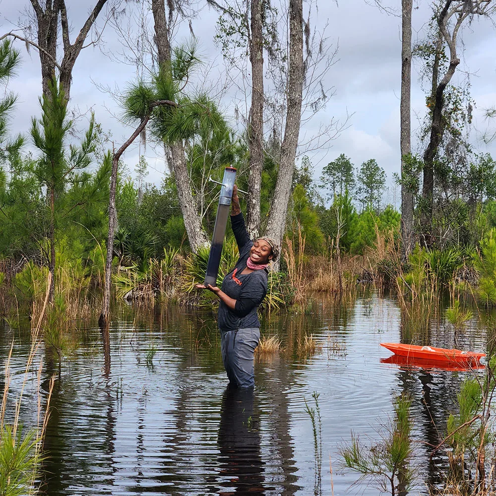 Jackson stands waist-deep in a wooded wetland, smiling as she holds a long sampling tube upright. An orange tub floats nearby.