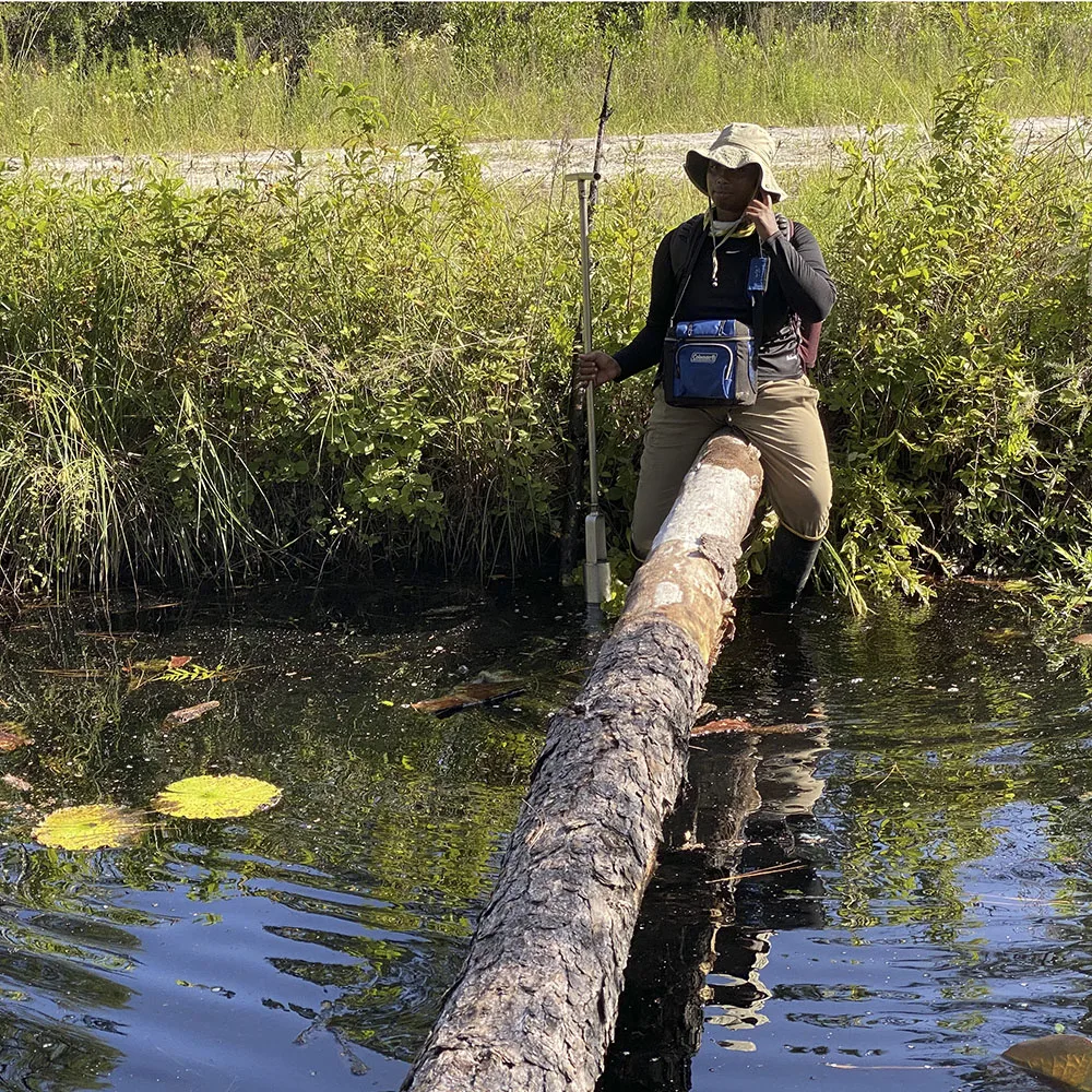 Alexis “AJ” Jackson stands in shallow water beside a fallen tree, holding a long sampling tool. Dense wetland vegetation lines the bank behind her.