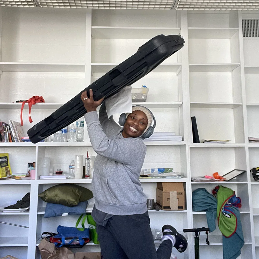Jackson smiles while lifting a long black equipment case over her shoulder in a room with white shelves.