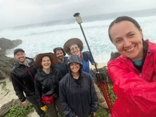 Six people in rain jackets pose for a selfie on a rocky coast, with waves breaking behind them.