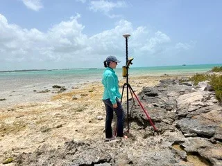 A person stands beside a tripod-mounted surveying instrument on a rocky shoreline, with shallow turquoise water in the background.